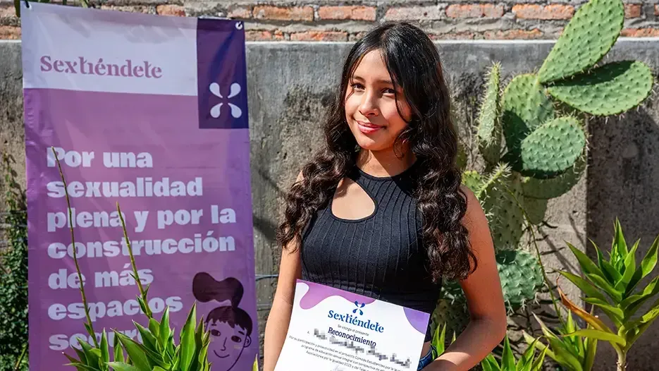 Estudiante con lentes posa con su reconocimiento frente a la lona morada