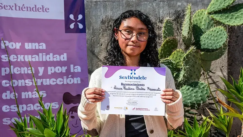 Estudiante de cabello rizado sostiene su reconocimiento junto a la lona de Sextiéndete