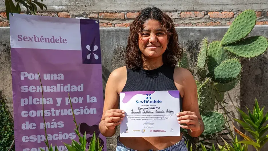 Estudiante con lentes muestra su reconocimiento frente a un nopal y la lona morada