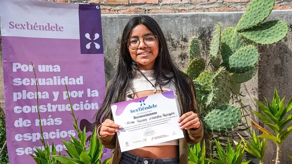 Estudiante de cabello largo sostiene su reconocimiento junto a la lona del programa