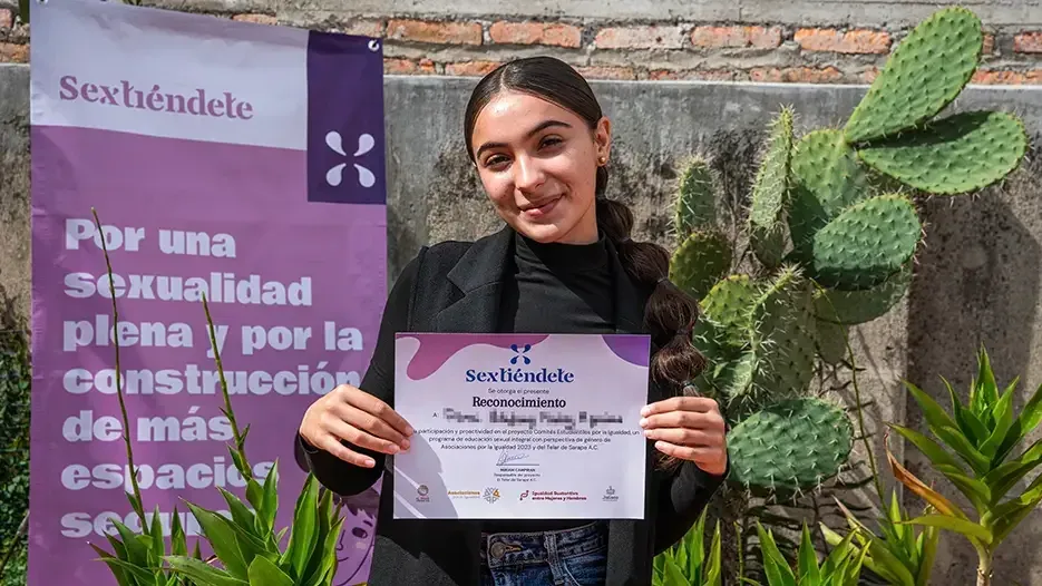 Estudiante sonríe con su reconocimiento frente a la lona morada de Sextiéndete