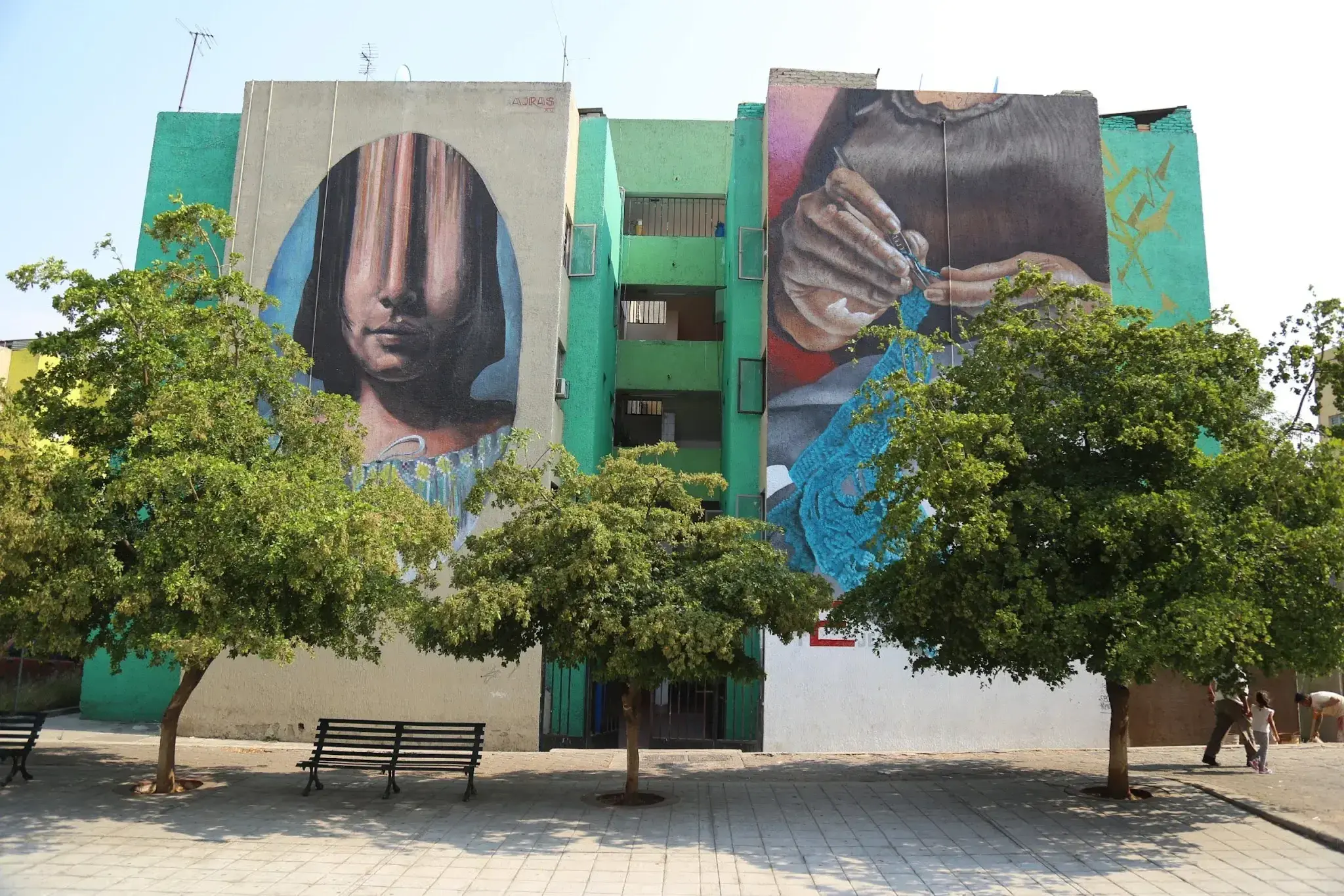 Mural de un rostro y manos pintado en un edificio verde, visto entre árboles de un parque