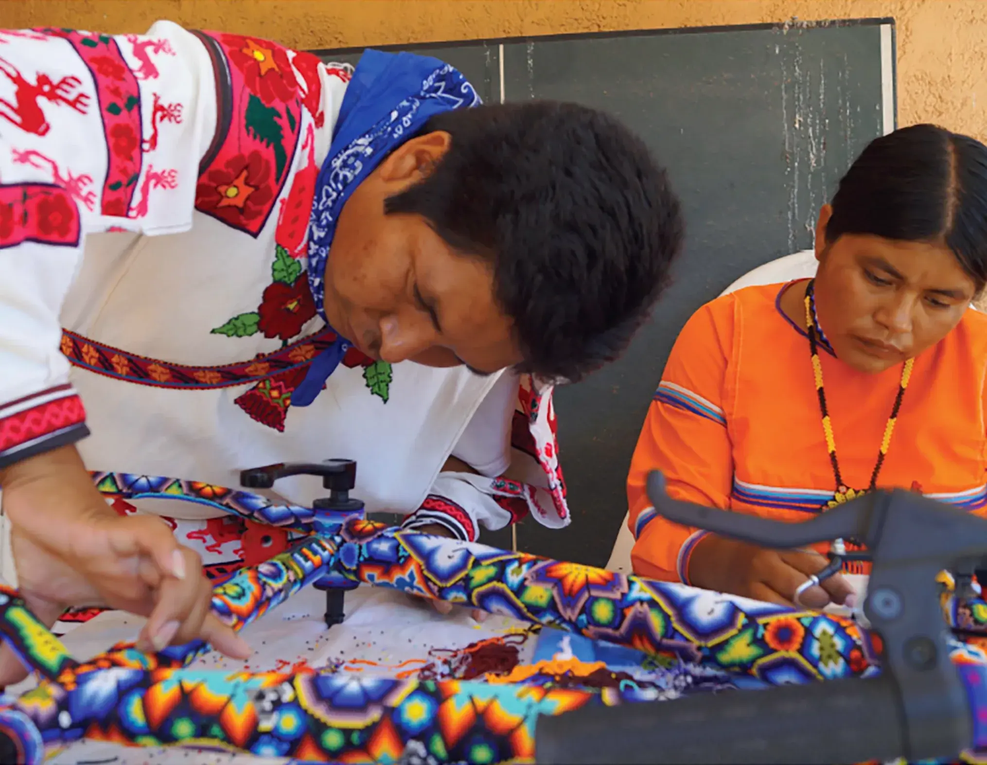 Artesano wixárika con camisa bordada decora una pieza con chaquira de colores junto a una mujer