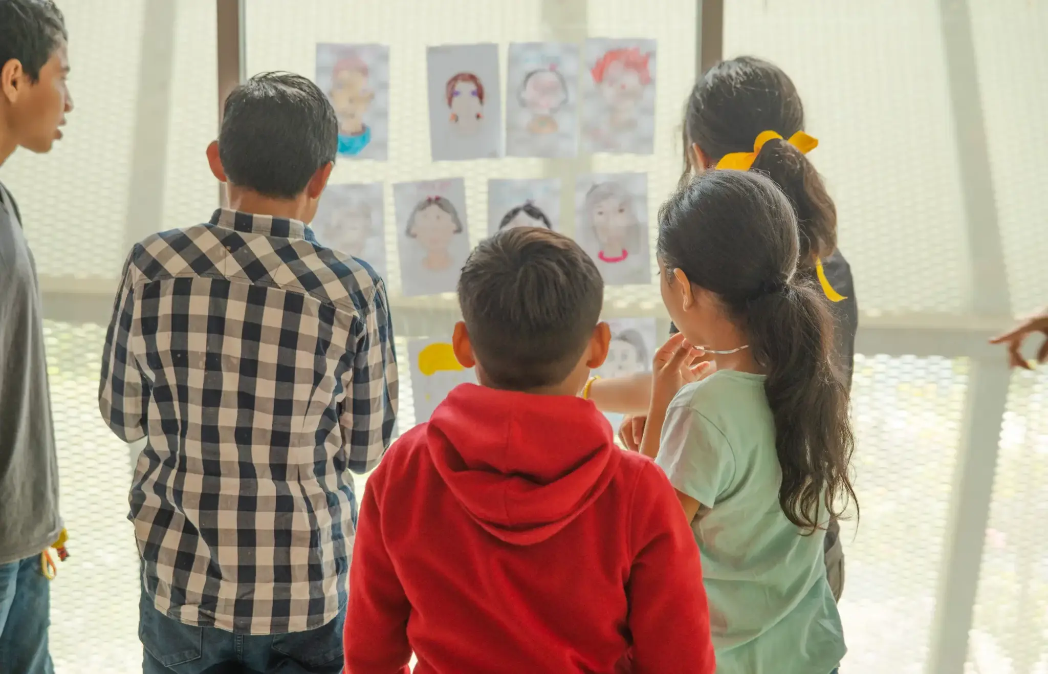 Niñas y niños observan de espaldas una pared con retratos dibujados