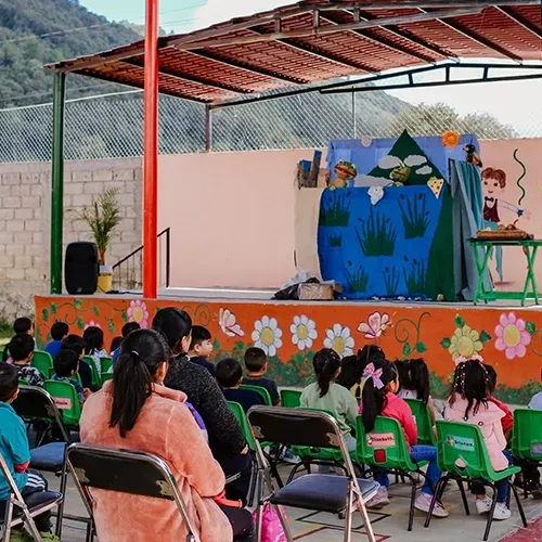 Niñas y niños sentados bajo un techo ven una obra de teatro con escenografía azul