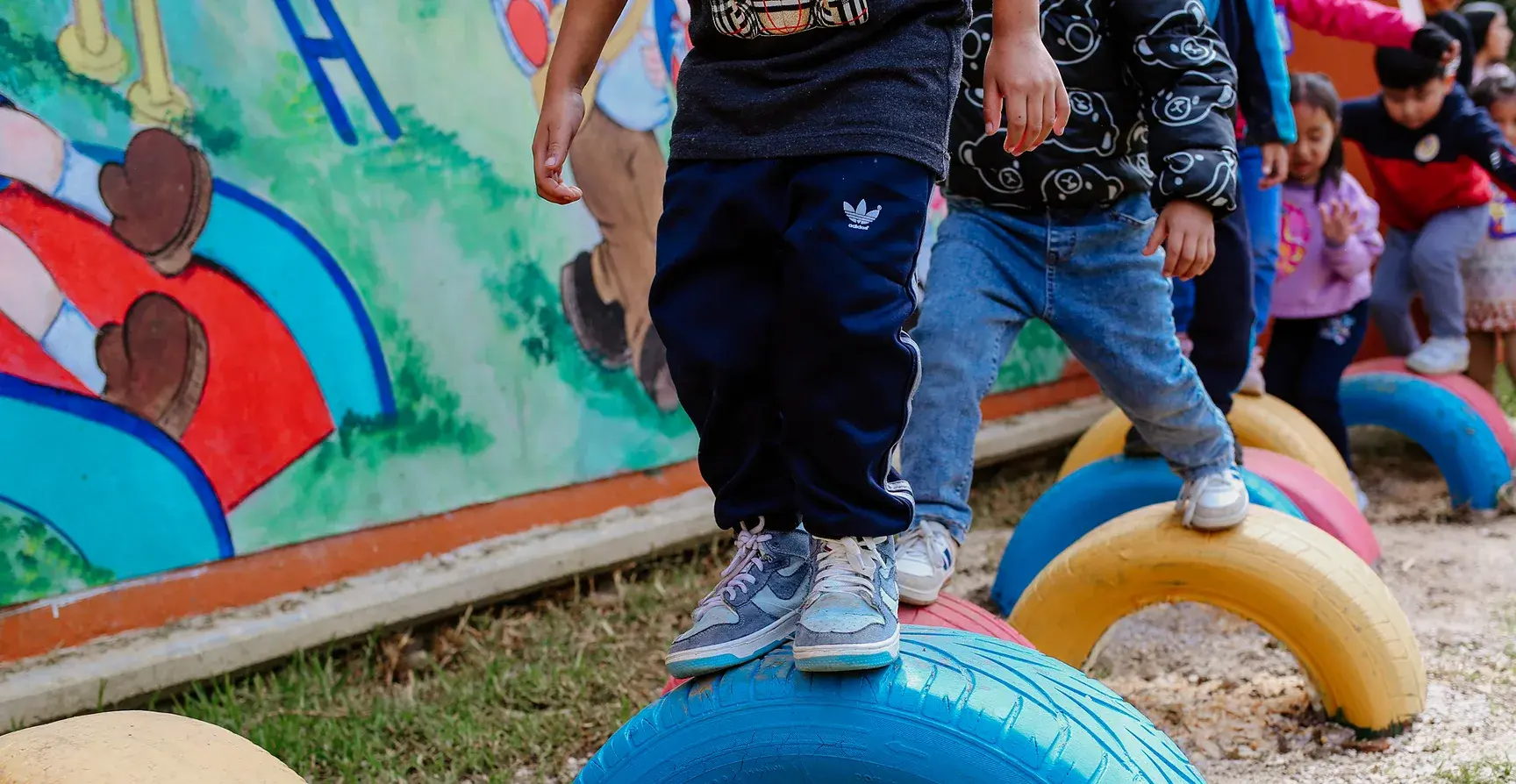 Niñas y niños caminan sobre llantas de colores frente a un mural en el patio escolar