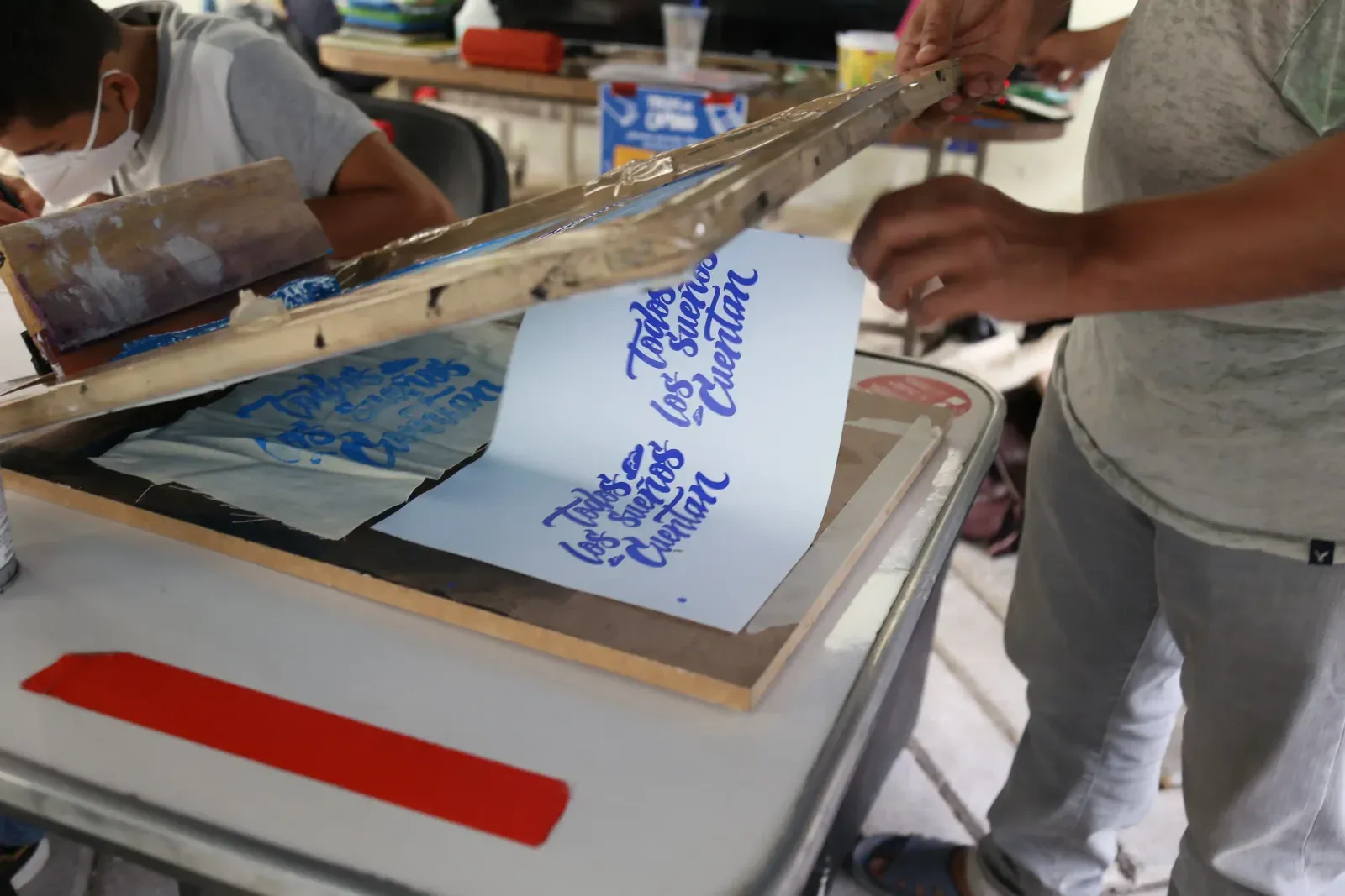 Participantes levantan una pantalla de serigrafía para revelar una impresión con lettering azul