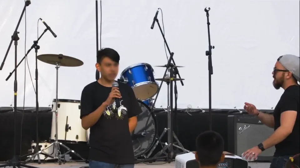 Joven con micrófono habla en un escenario con batería y equipo de sonido